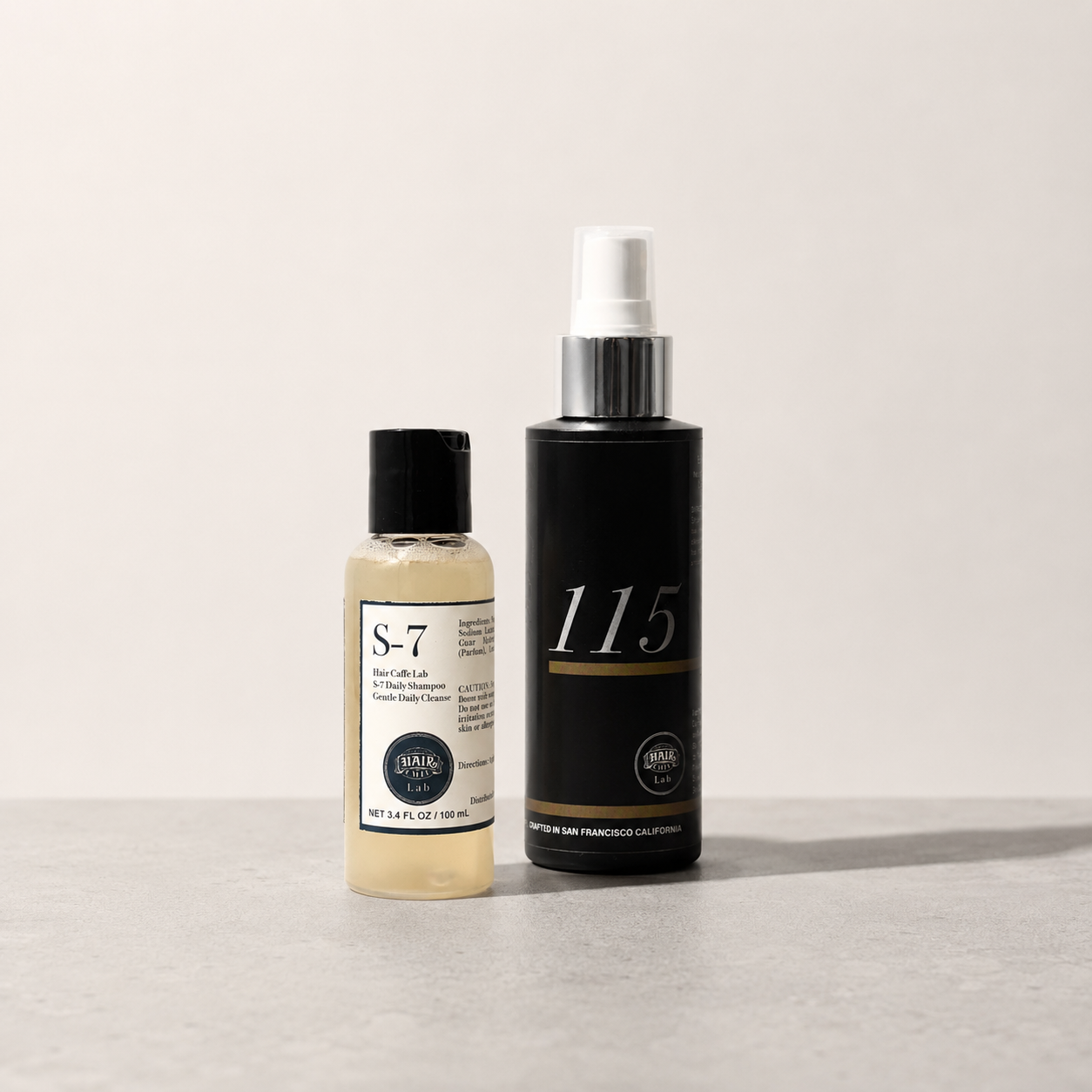 Hair Caffe Lab 2-Step Starter Set: S-7 Daily Shampoo 100 mL travel bottle beside the black No.115 Leave-In Treatment Mist, on a pale grey surface