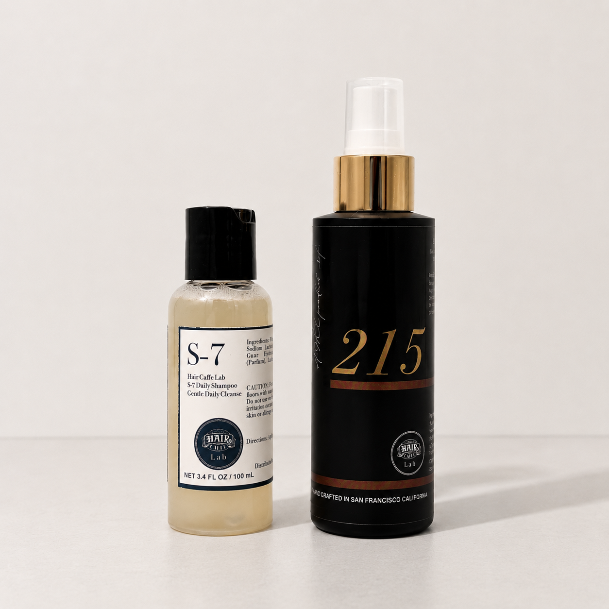 Hair Caffe Lab 2-Step Starter Set: S-7 Daily Shampoo 100 mL travel bottle beside the black No.215 Leave-In Treatment Mist with gold collar, on a pale grey surface